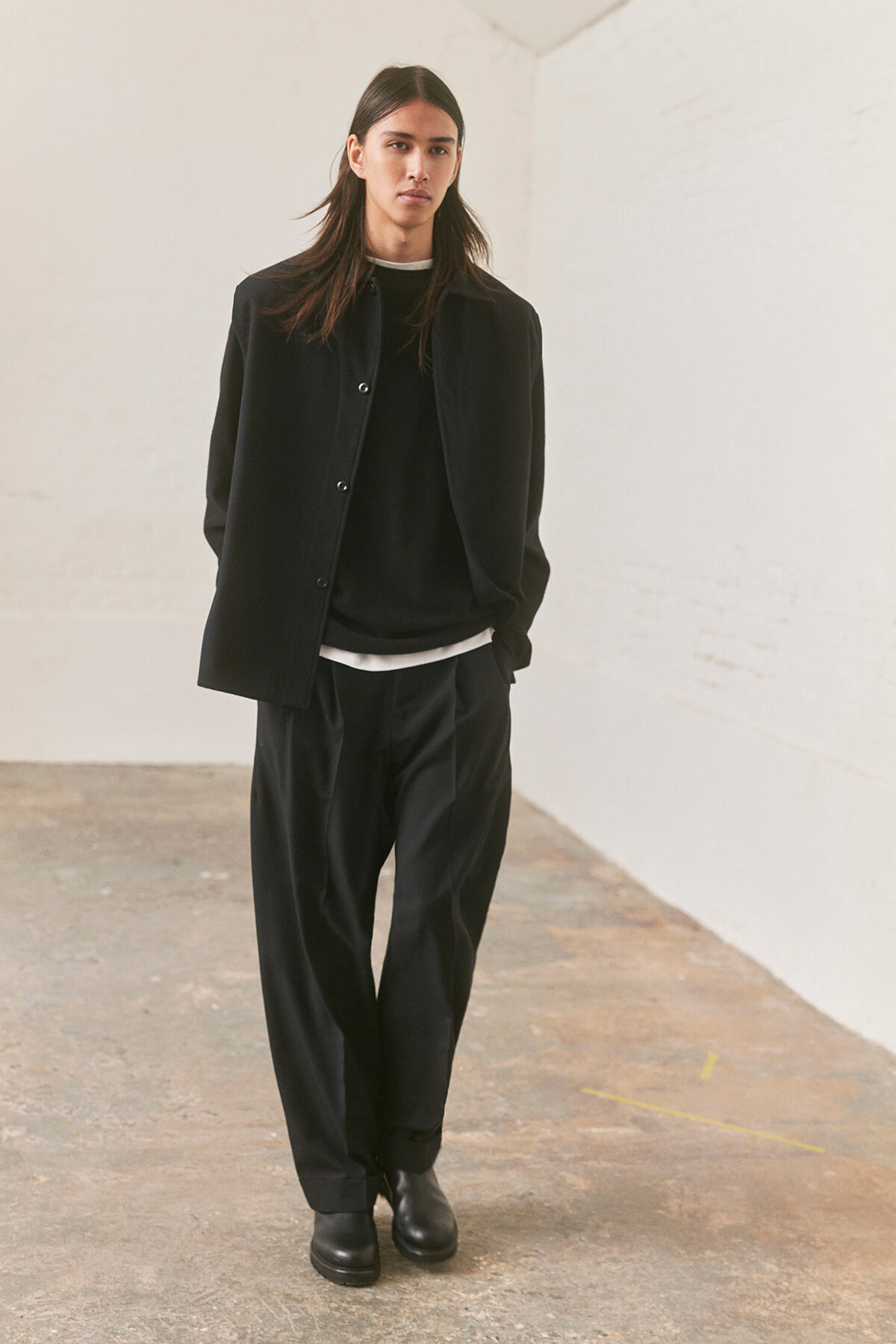 Model in an all-black outfit (coat, sweater, wide-leg trousers, boots) standing in a bright, minimalist room with white walls.