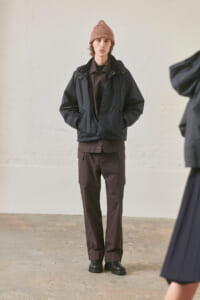 Young man standing indoors in a black jacket, brown cargo pants, black boots, and a pink knit beanie, with hands in pockets.