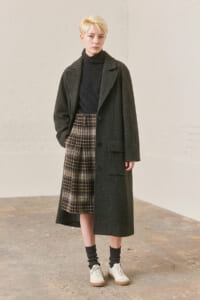 Model in a long dark overcoat, black turtleneck, and brown plaid midi skirt with white sneakers, standing in a neutral studio setting.