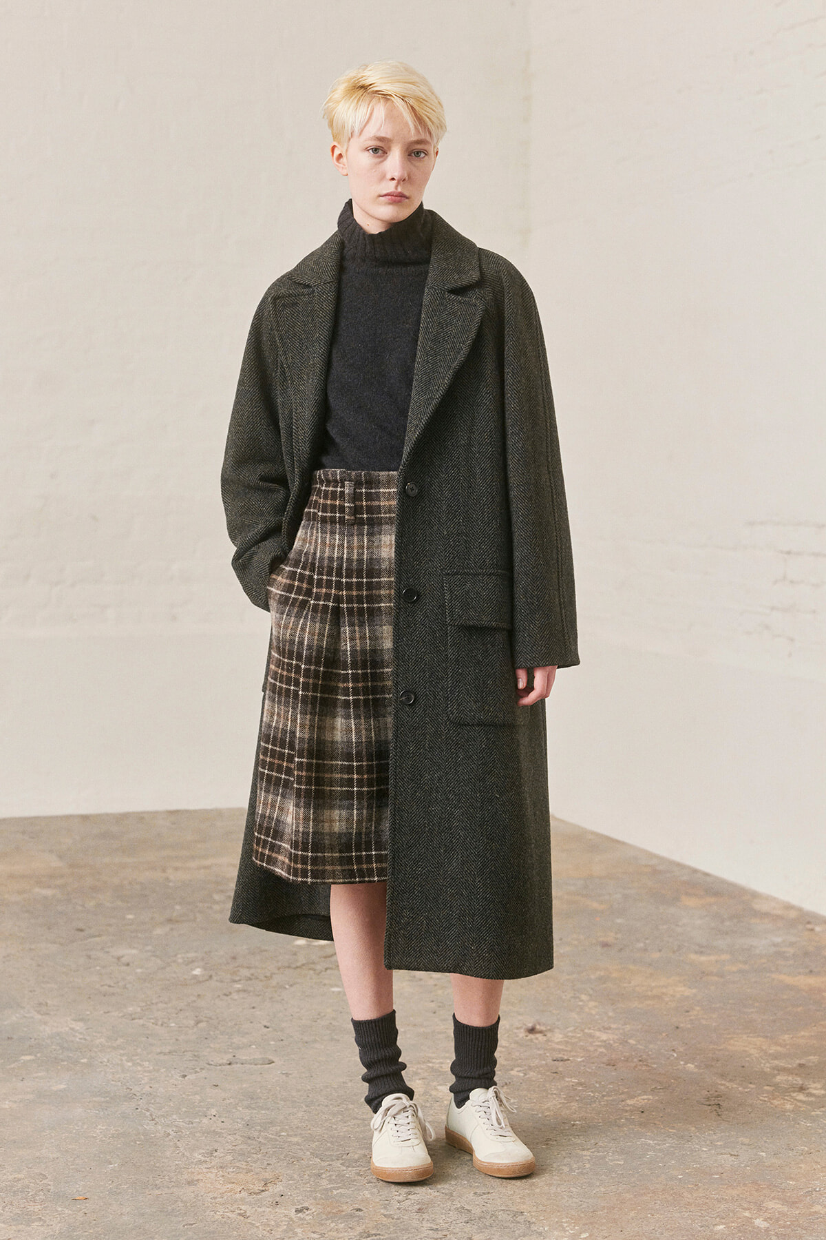 Model in a long dark overcoat, black turtleneck, and brown plaid midi skirt with white sneakers, standing in a neutral studio setting.
