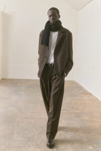 Man in a brown suit with a black scarf, white shirt, and belt stands in a neutral studio setting with hands in pockets.