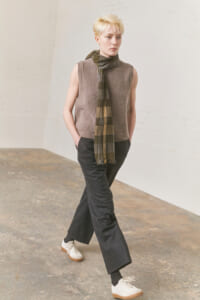 Full-body shot of a person wearing a taupe sleeveless knit top, plaid scarf, dark wide-leg pants, and white sneakers in a light studio/industrial space.