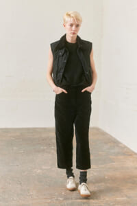 Person with short blond hair stands in a studio, wearing a black sleeveless vest, black knit top, black wide-leg pants, gray socks, and white sneakers.