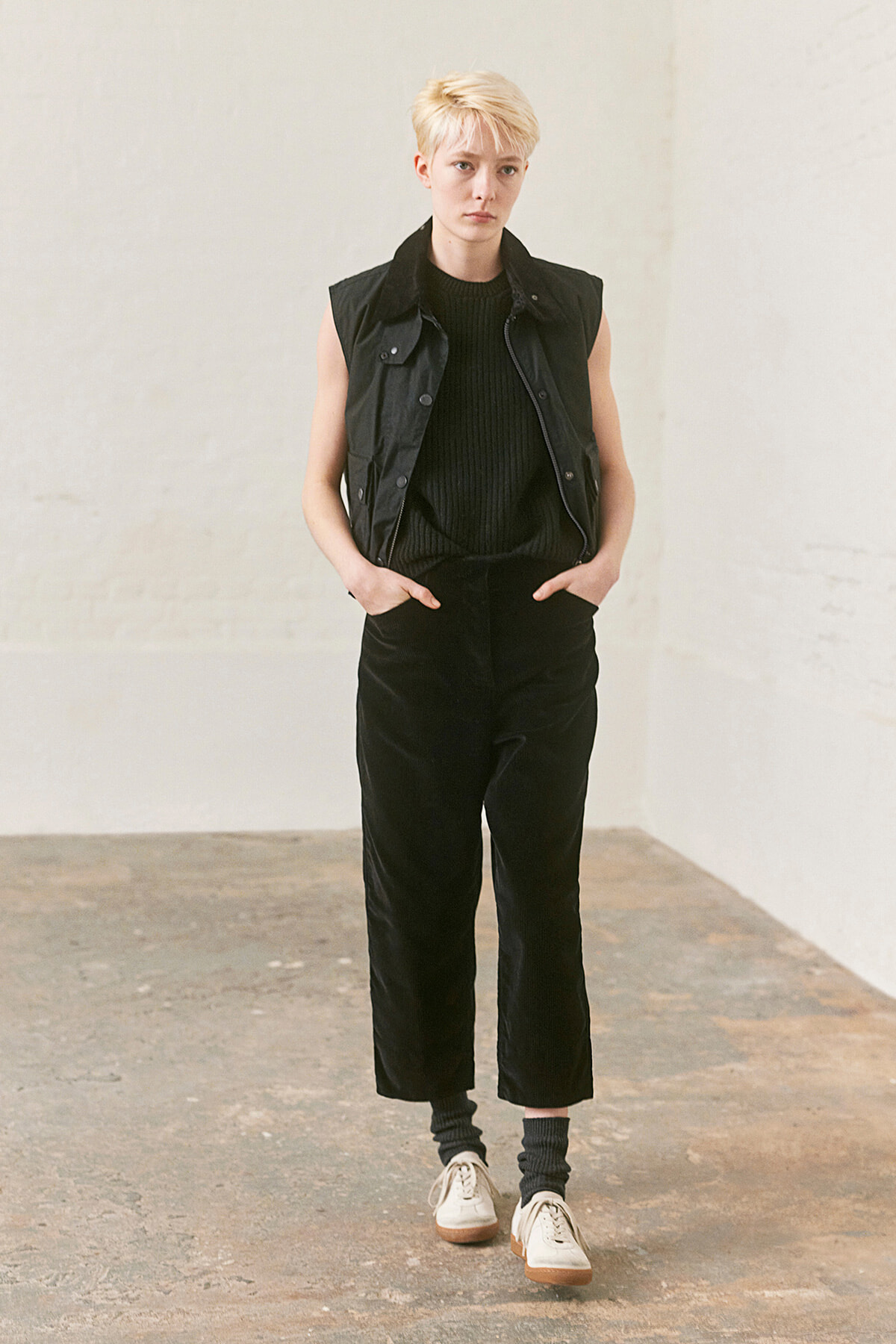 Person with short blond hair stands in a studio, wearing a black sleeveless vest, black knit top, black wide-leg pants, gray socks, and white sneakers.