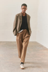 Model wearing a brown plaid blazer over a black sweater and white shirt, with tan pleated trousers and beige sneakers in a neutral studio setting