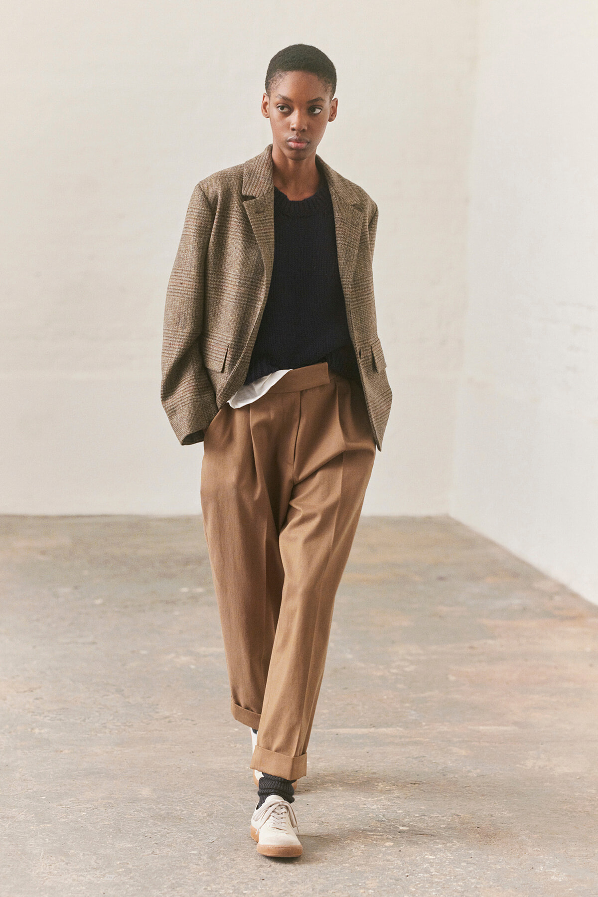 Model wearing a brown plaid blazer over a black sweater and white shirt, with tan pleated trousers and beige sneakers in a neutral studio setting