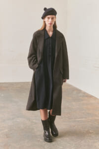 Model wearing a long dark wool coat over a navy dress with black boots and a navy beret in a minimalist studio setting.