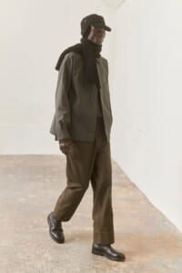 Person wearing an olive jacket, black scarf and cap, walking along a light-walled space.