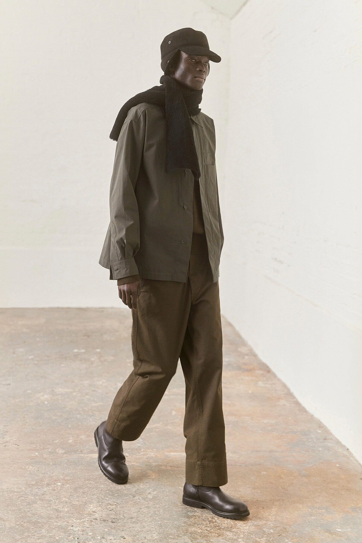 Person wearing an olive jacket, black scarf and cap, walking along a light-walled space.