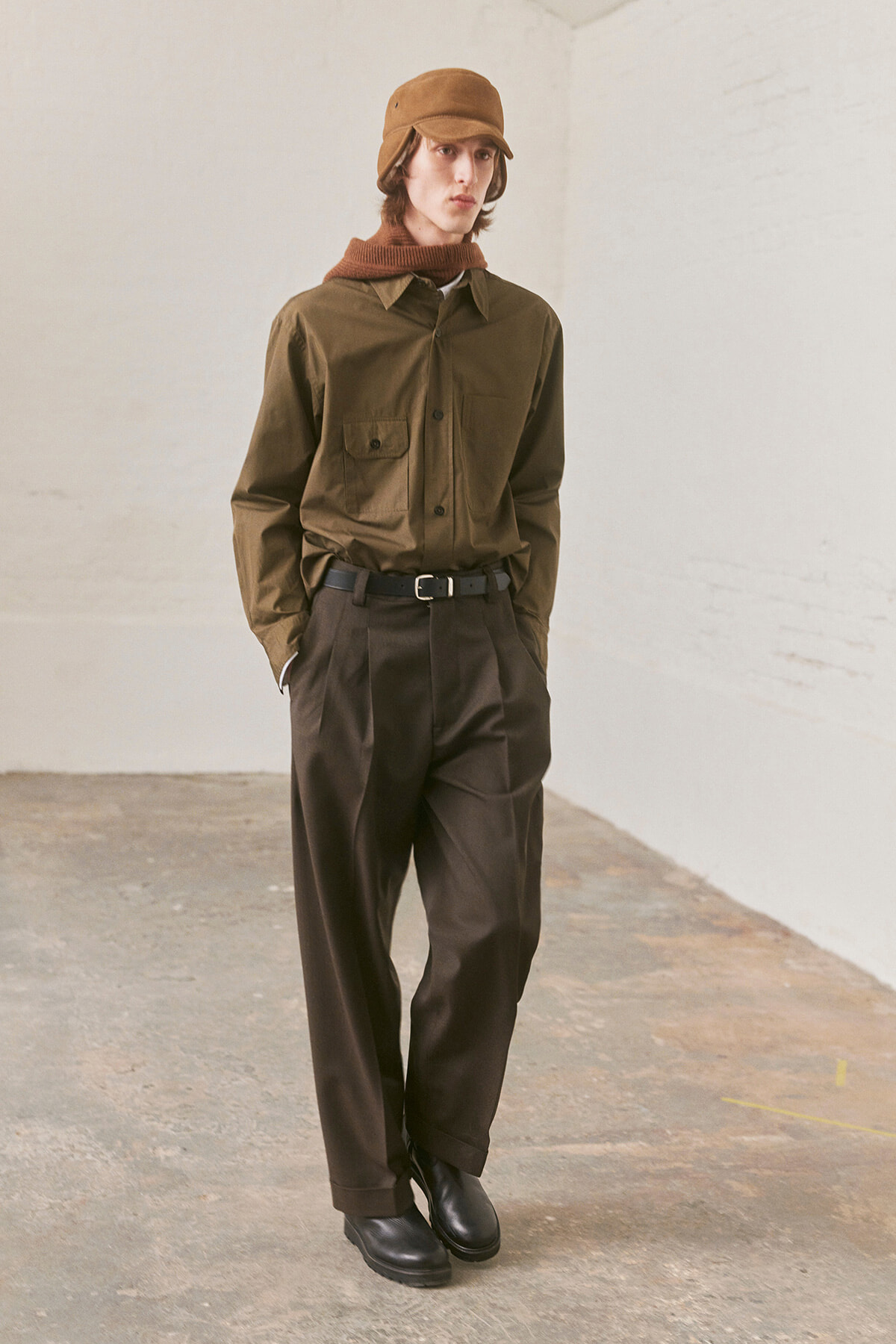 Man model in olive utility shirt, dark brown wide-leg trousers, brown belt, black shoes, and a brown cap in a minimal studio.