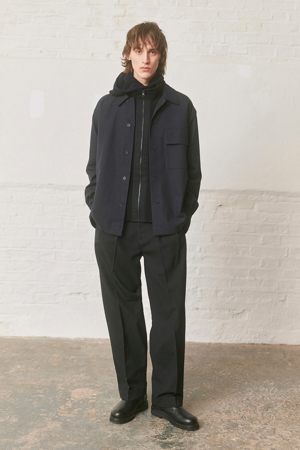 Young man in a dark navy jacket over a black hoodie, black pants, and black boots, standing with hands in pockets against a white brick wall.