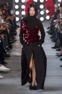 Model on a runway wearing a long black coat with a burgundy sequin appliqué top and a high slit in the skirt, audience in the background.