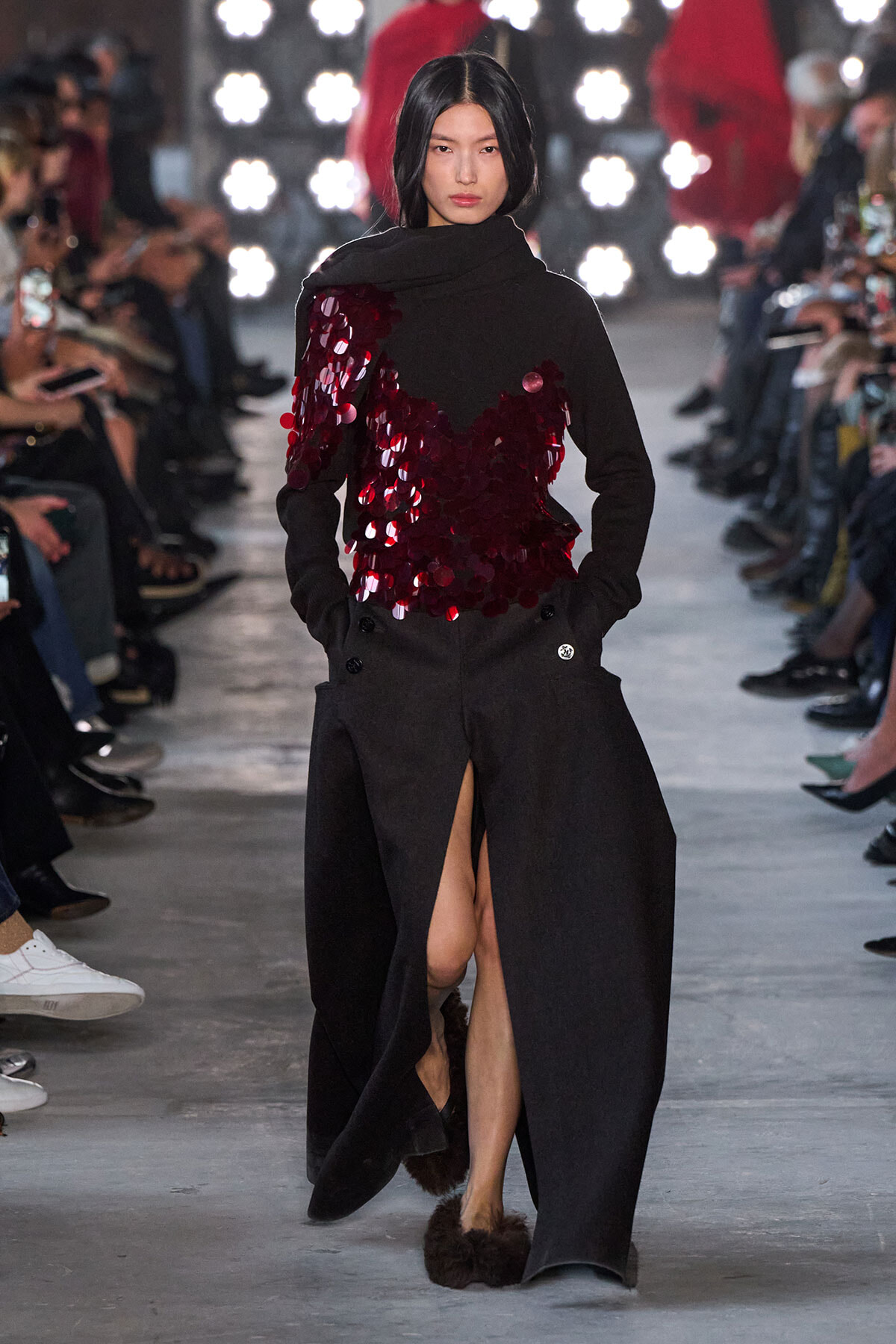 Model on a runway wearing a long black coat with a burgundy sequin appliqué top and a high slit in the skirt, audience in the background.