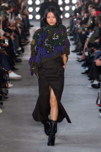 Model walks the runway wearing a fringed olive sweater with purple and green tassels, paired with a brown skirt and black boots.