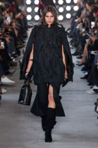 Model walking a runway in a black feathered cape-style dress with a front slit, paired with black gloves, boots, and a black handbag.