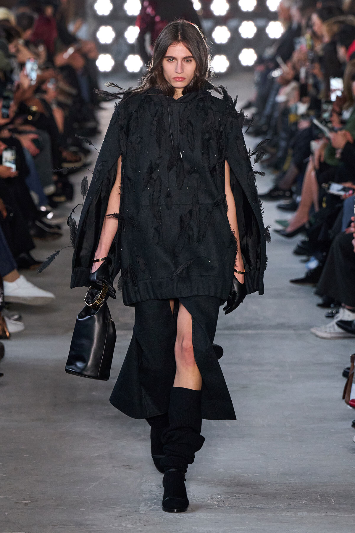 Model walking a runway in a black feathered cape-style dress with a front slit, paired with black gloves, boots, and a black handbag.