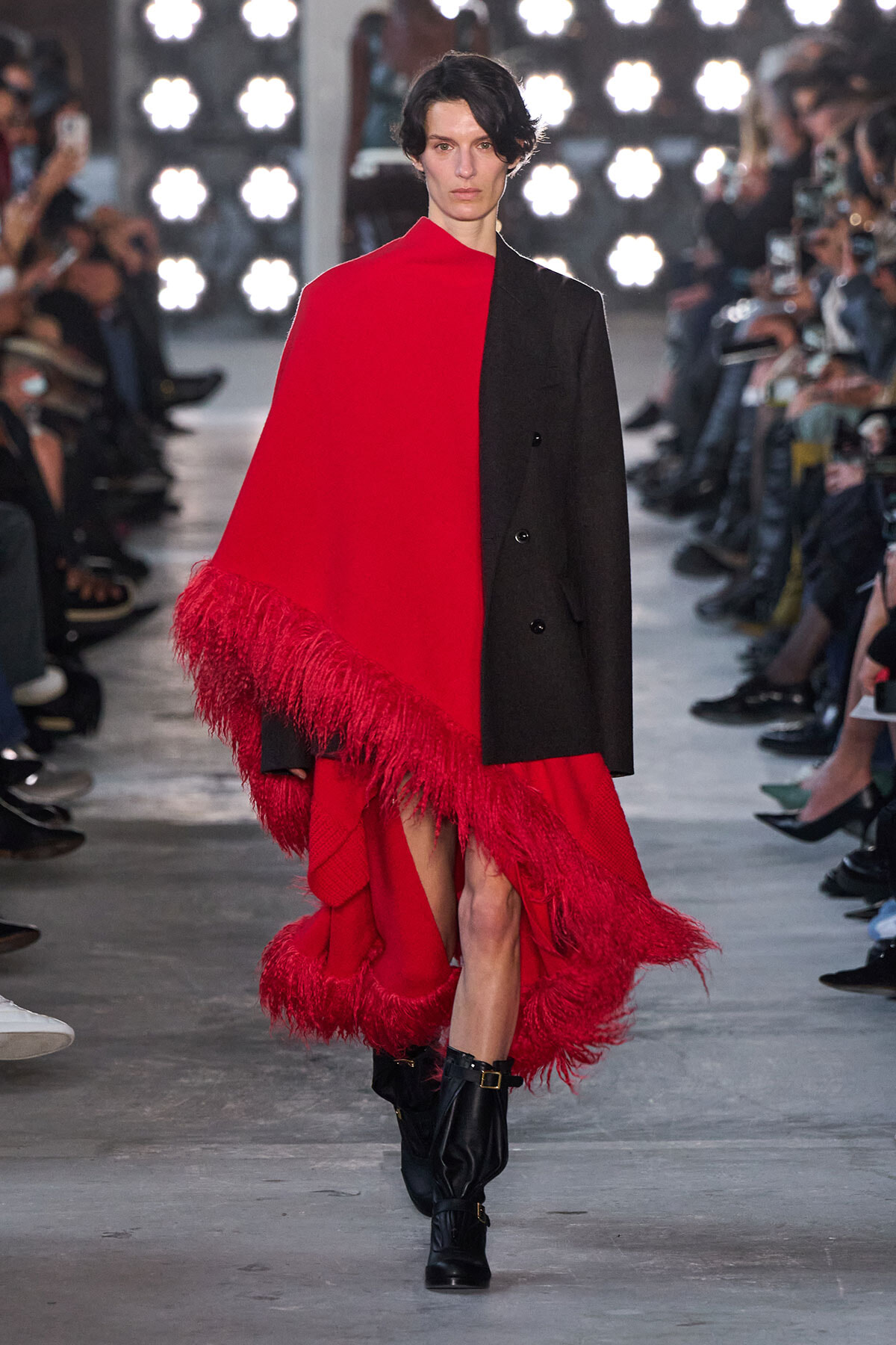 Model walking a runway in a bold red cape with fringe over a black blazer and black boots, audience in background (fashion show).