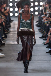 Model on a fashion runway wearing a teal leather bustier and an asymmetrical brown leather wrap skirt with a front zipper, fur waistband, black gloves, and knee boots.