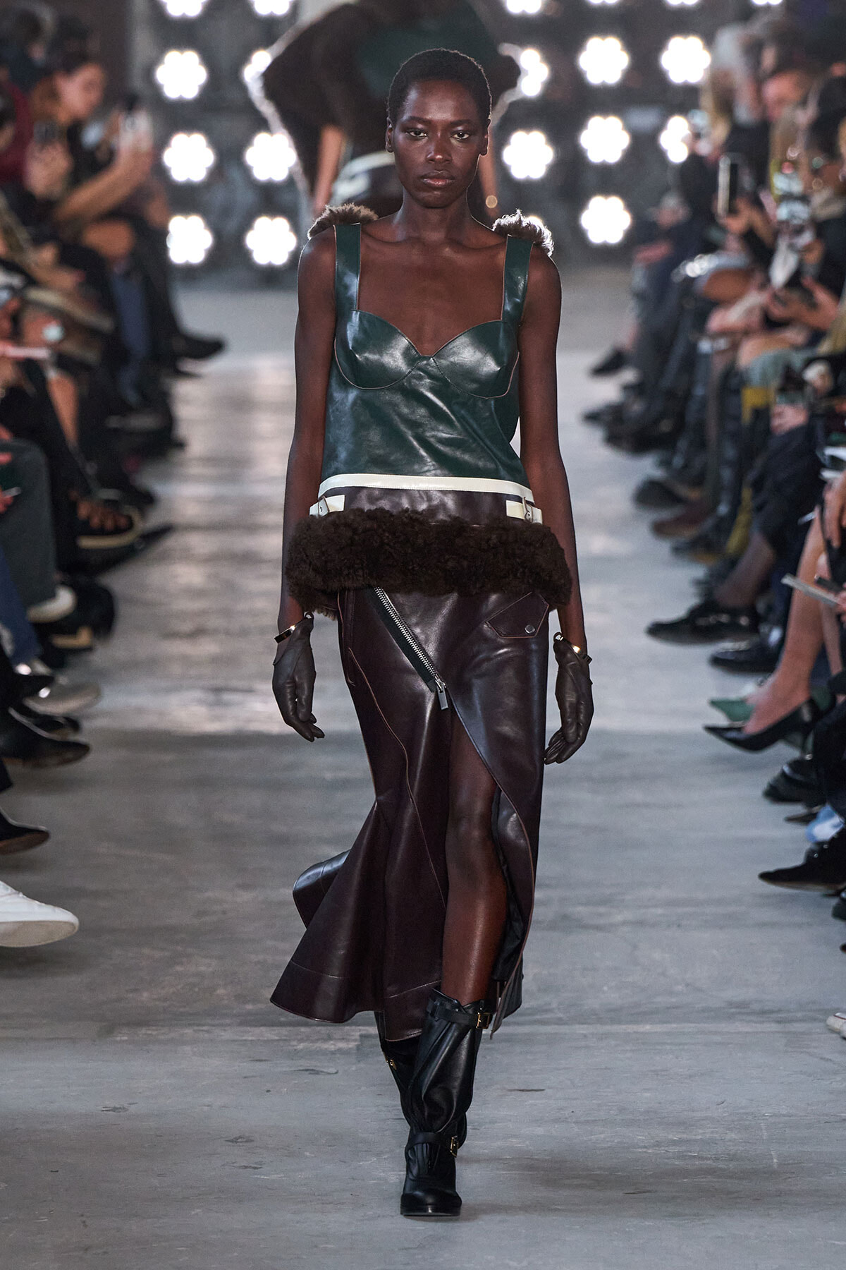 Model on a fashion runway wearing a teal leather bustier and an asymmetrical brown leather wrap skirt with a front zipper, fur waistband, black gloves, and knee boots.