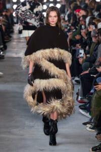 Model walking a runway in a black cape-like garment with cream feather fringe spiraling around her torso; audience watches in the background.