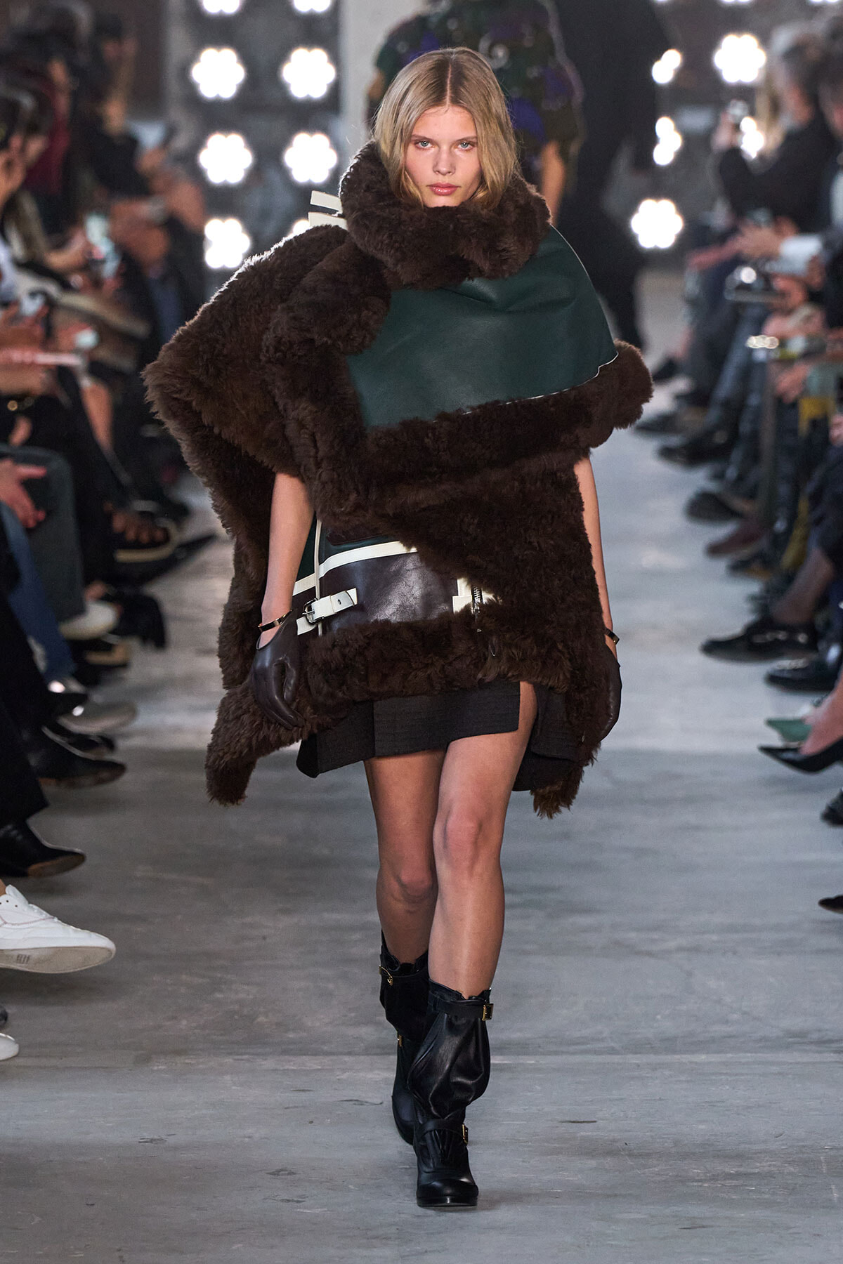 Model walks the runway wearing an oversized brown shearling coat with a green panel, a black mini skirt, gloves, and black boots.