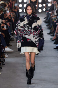 Model walks down runway in oversized fringed black, white, and red patterned poncho over a white mini skirt, black boots; audience in background.