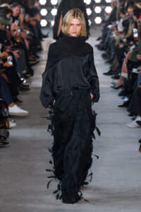 Model walking a fashion runway in a black, draped top and voluminous feathered trousers, hands in pockets, audience blurred in the background.
