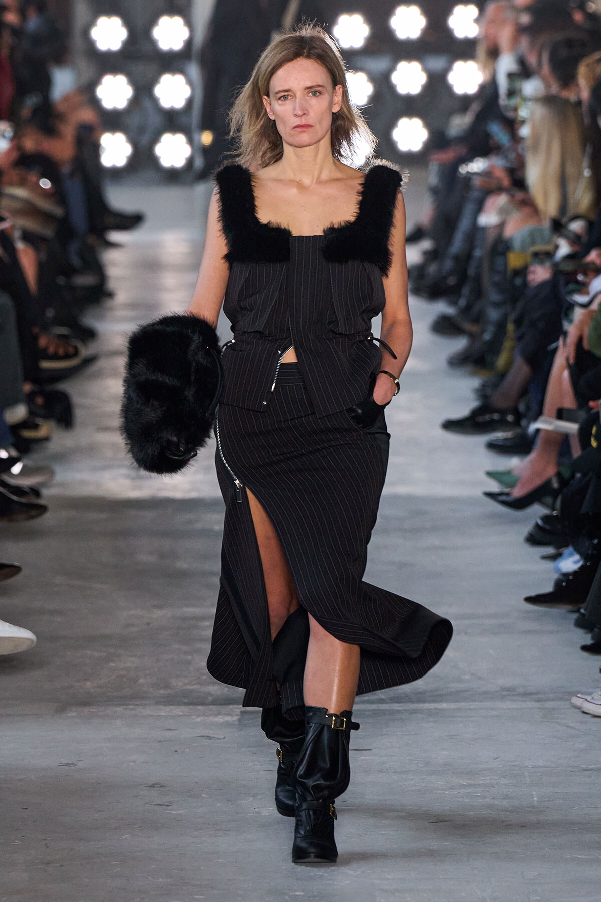 Model on a runway wearing a black pinstriped dress with fur trim, carrying a black fur bag and wearing black ankle boots.
