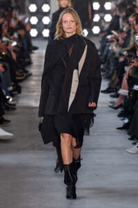 Model walks a fashion runway in a oversized pinstripe black blazer with layered panels and a sheer inner layer, black boots nearby.