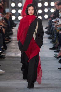Model walking a fashion show runway in an oversized black blazer paired with a bold red sheer scarf draped over one shoulder and a flowing red overlay, with wide-leg black pants; bright stage lights and seated audience along the sides.