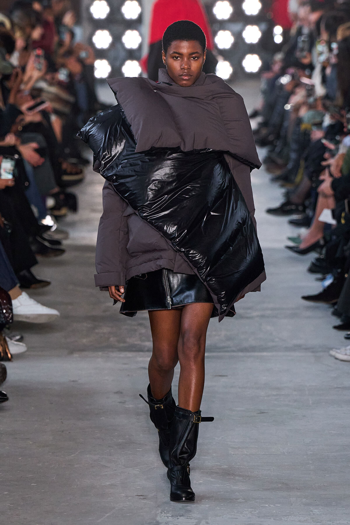 Model walking the runway in an oversized taupe puffer with a large black glossy panel, paired with black boots.