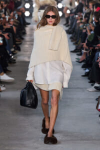 Model walks runway in oversized cream knit poncho over white shirt dress, black sunglasses, and brown furry slippers, carrying a black handbag.