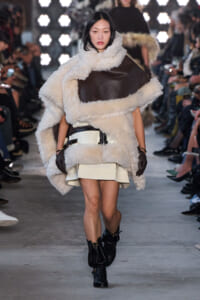 Model walks the runway in an oversized cream shearling coat with brown leather panels, paired with a cream mini skirt, black gloves, and black buckled boots.