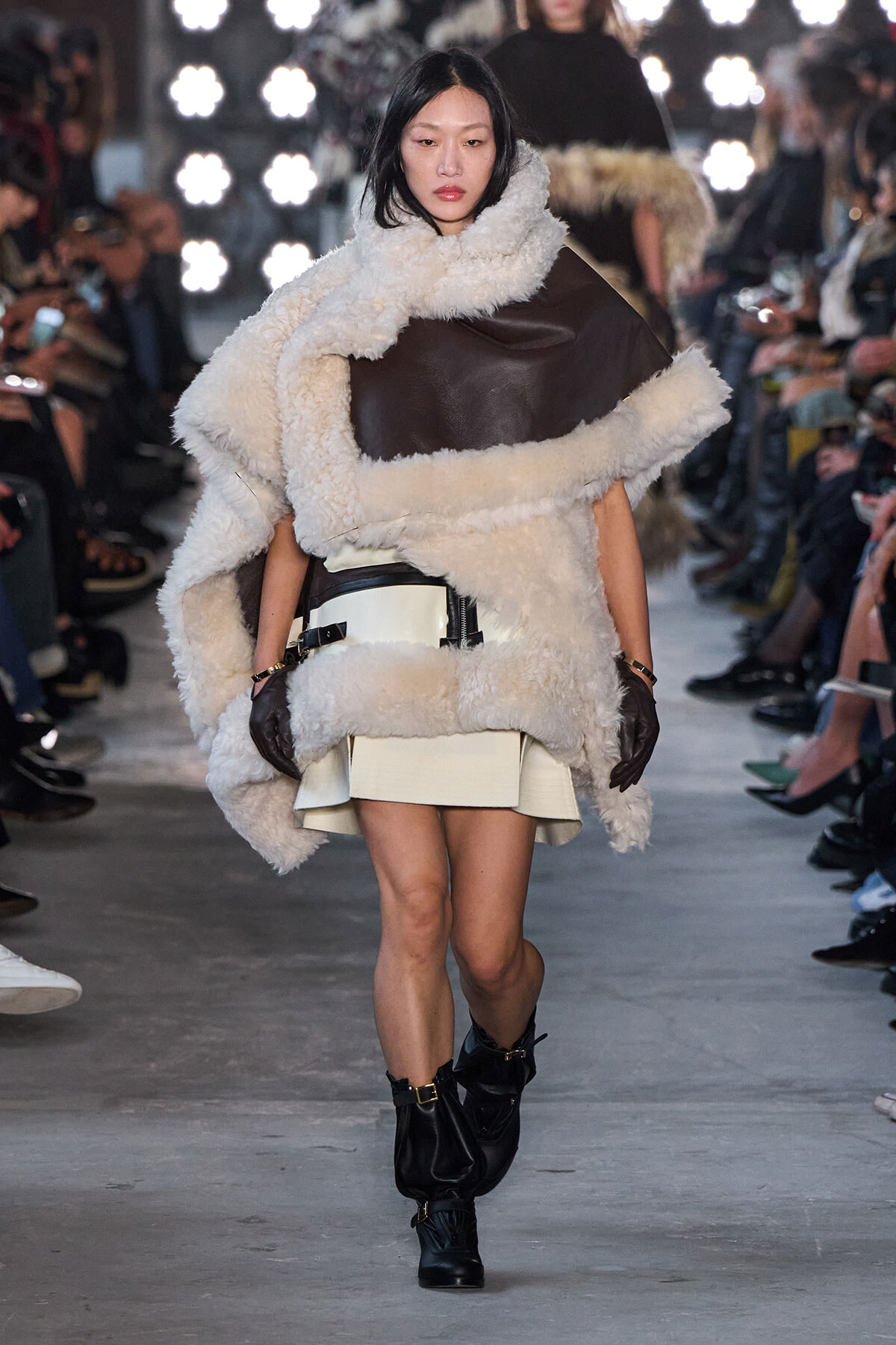 Model walks the runway in an oversized cream shearling coat with brown leather panels, paired with a cream mini skirt, black gloves, and black buckled boots.