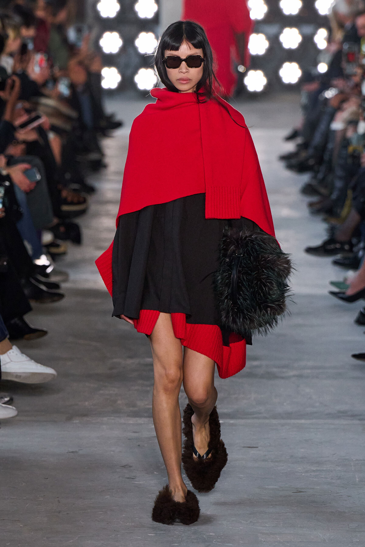 Model walking runway in a bold red cape layered over a black dress, wearing dark sunglasses and furry slippers, holding a black furry bag with her left hand.