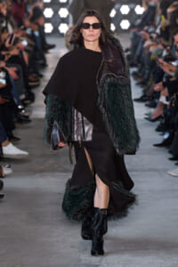 Model walking a runway in a black ensemble with green fur-trimmed sleeves and sunglasses, audience on both sides.