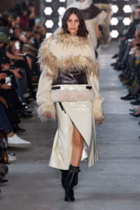 Model walks a runway in a cream fur-trimmed coat with a wide leather belt and black boots, audience blurred in the background