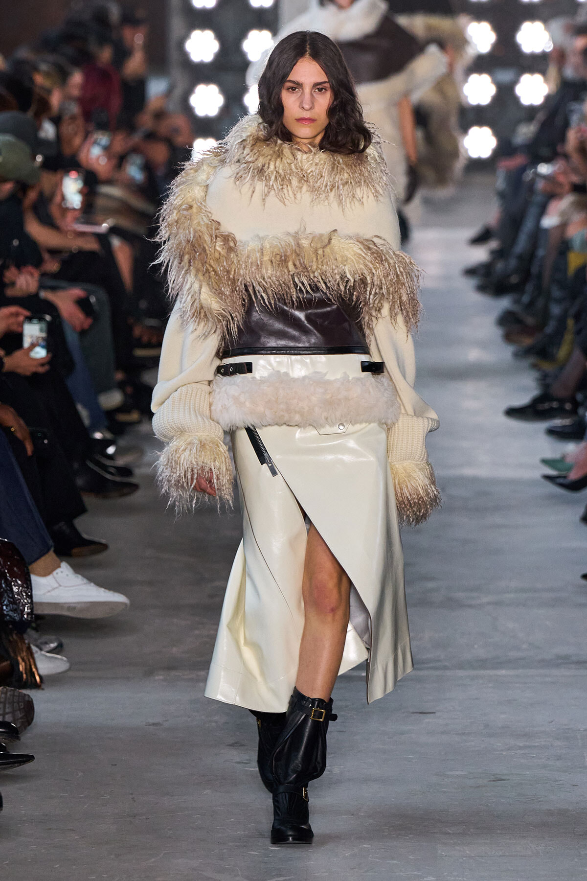 Model walks a runway in a cream fur-trimmed coat with a wide leather belt and black boots, audience blurred in the background