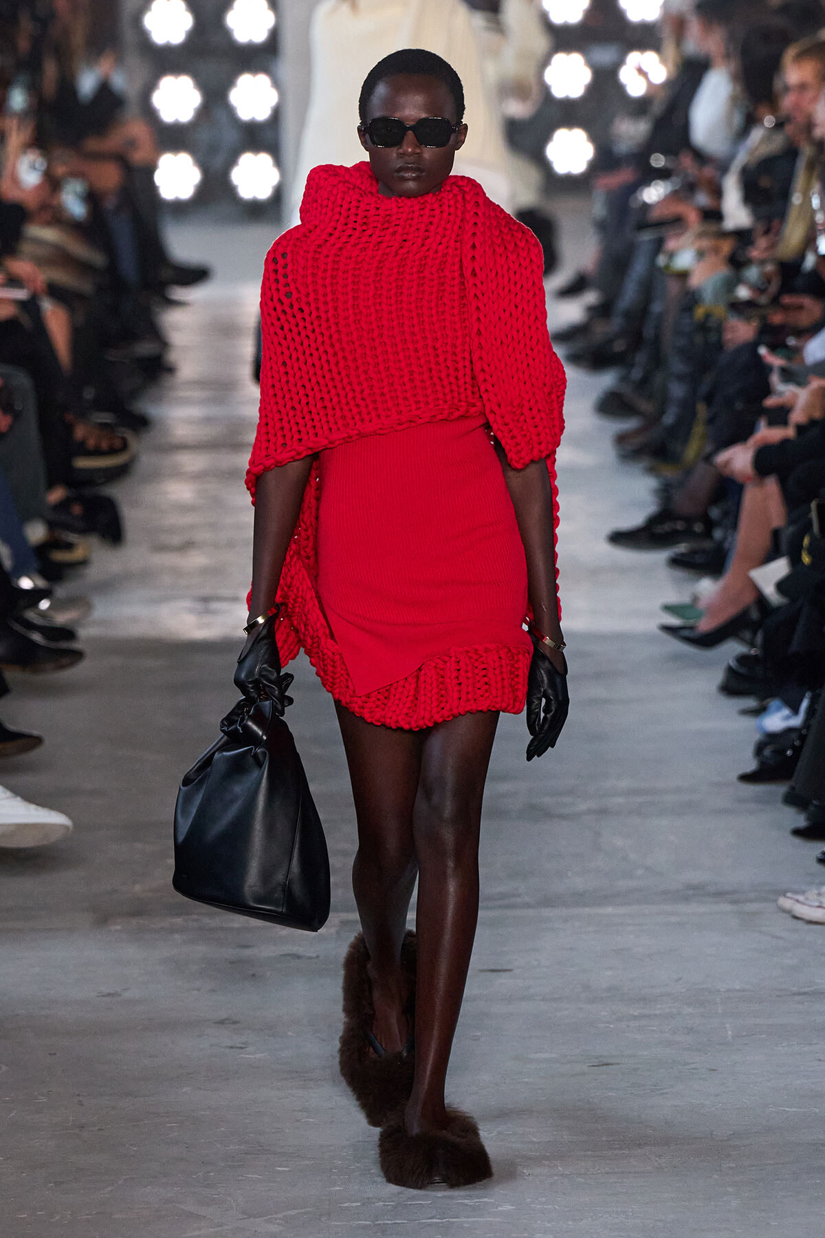 Model walking a runway in a bright red chunky-knit cape over a bodycon dress, with black sunglasses, gloves, a large black bag, and fur slippers.
