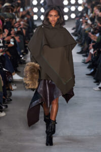 Runway model wearing an oversized olive cape with an asymmetric hem, a brown leather skirt, black buckled boots, and a fur handbag on the runway.