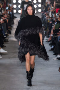 Female model walking a runway in a black cape with shaggy feathered fringe, paired with black knee-high boots; audience lines the catwalk in the background.