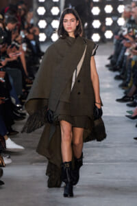 Model walking a runway in an oversized olive-green cape coat with fringe at the hem, black gloves, and ankle boots, as photographers line the aisle.