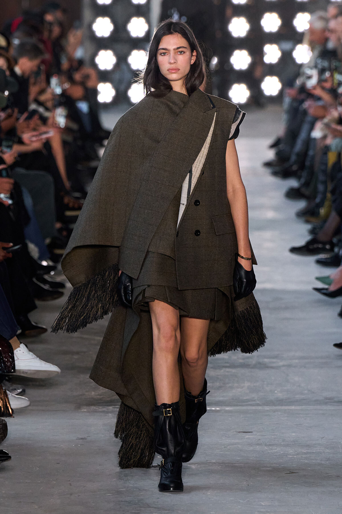 Model walking a runway in an oversized olive-green cape coat with fringe at the hem, black gloves, and ankle boots, as photographers line the aisle.