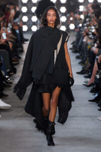 Model walking a runway in a dark layered cape with fringe and asymmetrical panels, gloves, and ankle boots.