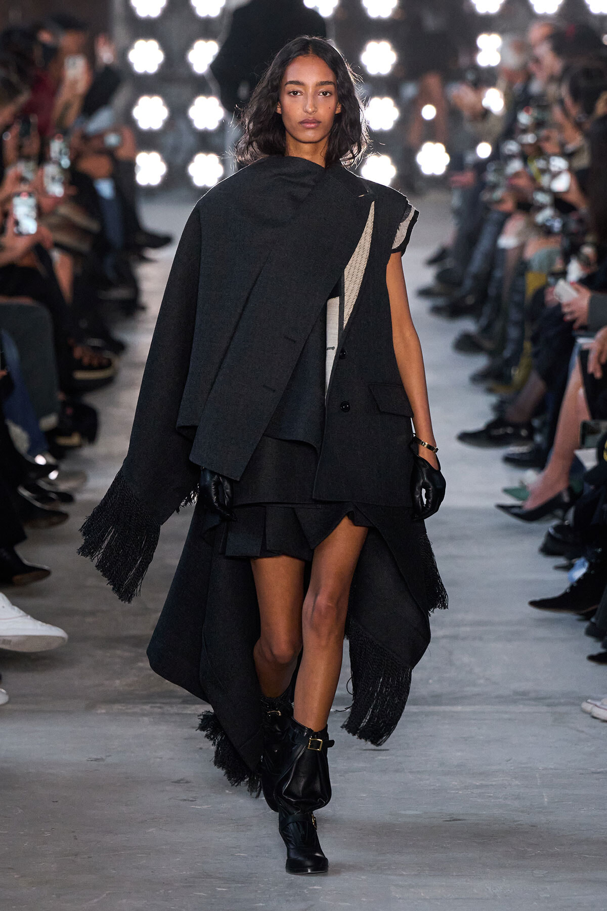 Model walking a runway in a dark layered cape with fringe and asymmetrical panels, gloves, and ankle boots.