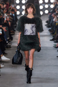 Runway model in a fuzzy green mini dress with a grayscale portrait panel, holding a black handbag and wearing buckled boots.