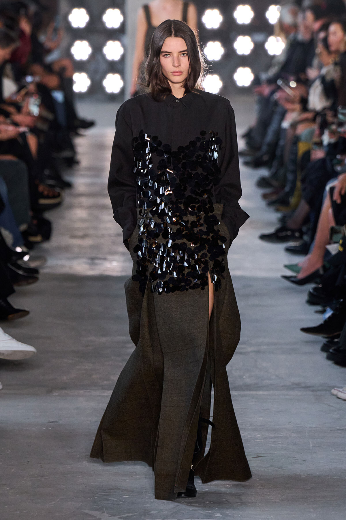 Model walking a runway in a black blouse with a sequined front and a long brown skirt with a slit, audience on both sides.