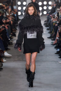 Model on a runway in a black knit mini dress with feathered trim at the collar and cuffs, wearing black boots.