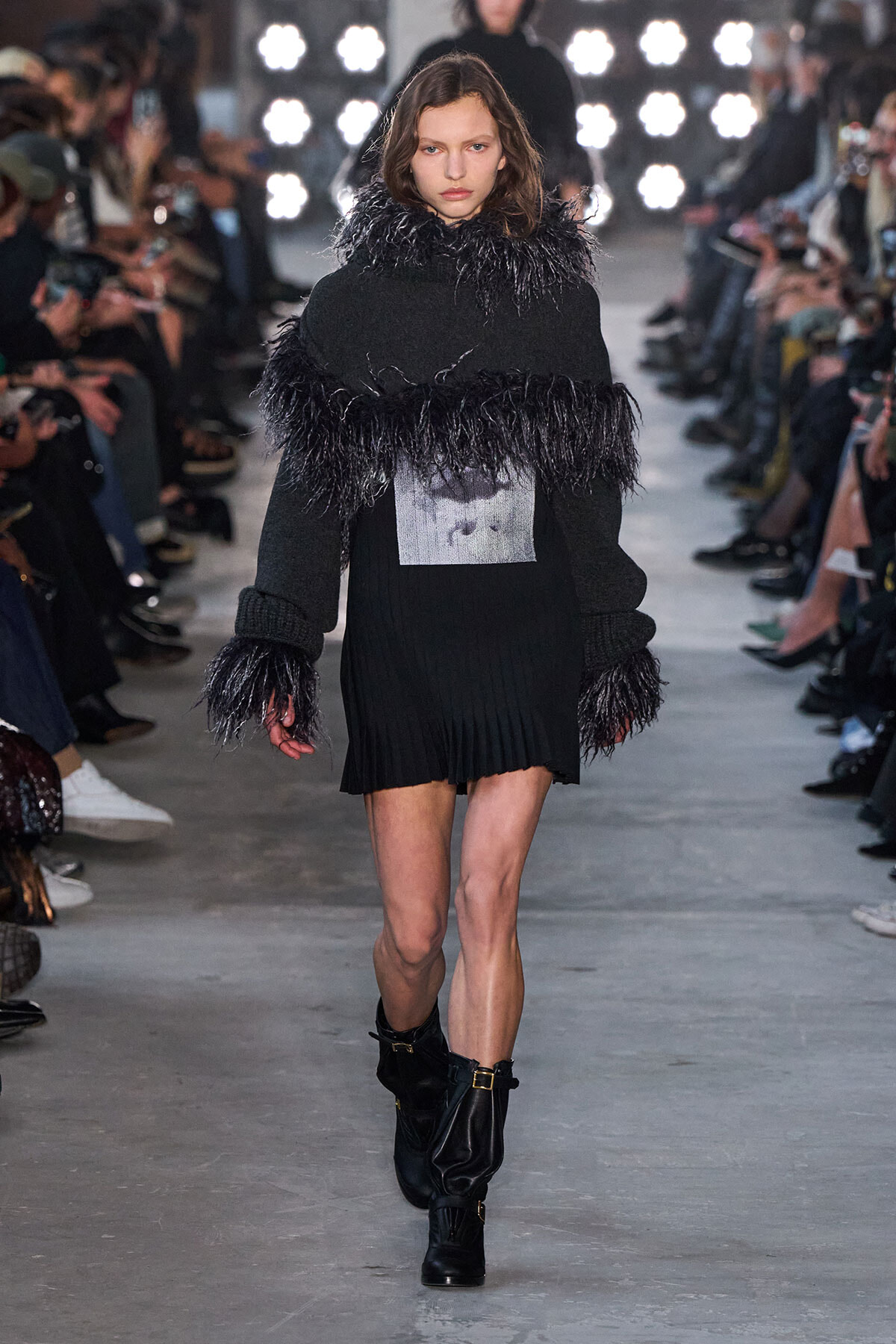 Model on a runway in a black knit mini dress with feathered trim at the collar and cuffs, wearing black boots.