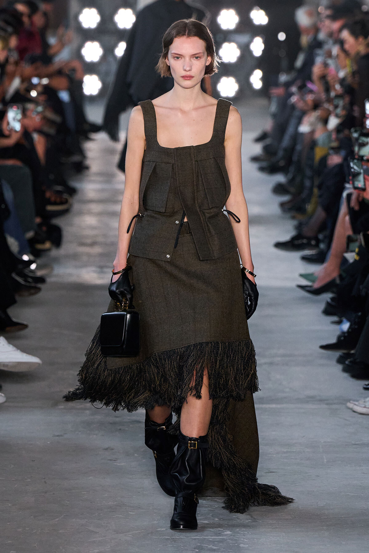 Model walks a runway in a brown textured dress with a fringe hem and matching vest, paired with black gloves and boots, carrying a small black box handbag.